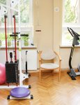 Wnętrze domu pomocy. Gabinet rehabilitacji ruchowej. Na drewnianej podłodze stoją duży wielofunkcyjny atlas do ćwiczeń i rowerek treningowy. W tle duże okna z widokiem na ogród